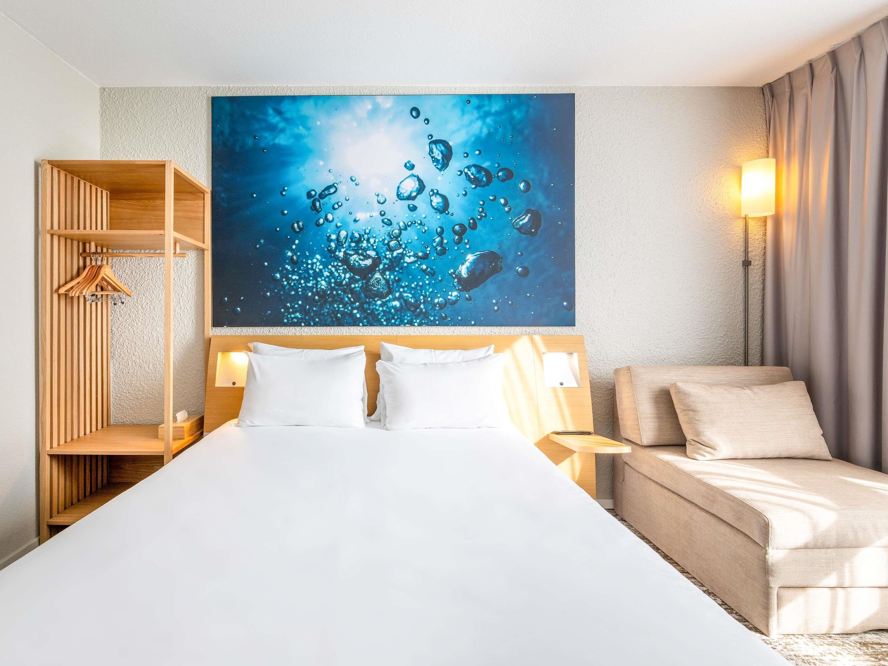 Una habitación de hotel con una cama, un armario de madera, un cuadro colgado en la pared y una mesita de noche con lámpara.