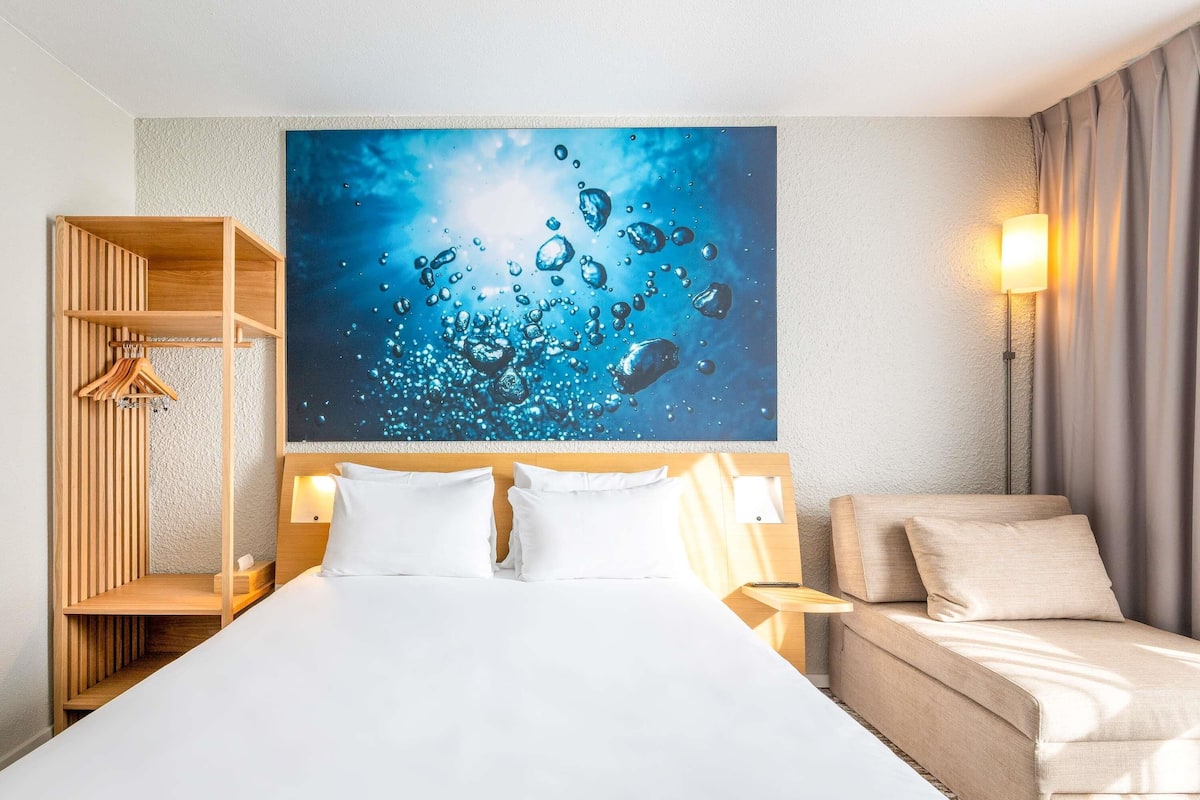 A hotel room with a bed, a wooden wardrobe, a wall-mounted painting, and a bedside table with a lamp.
