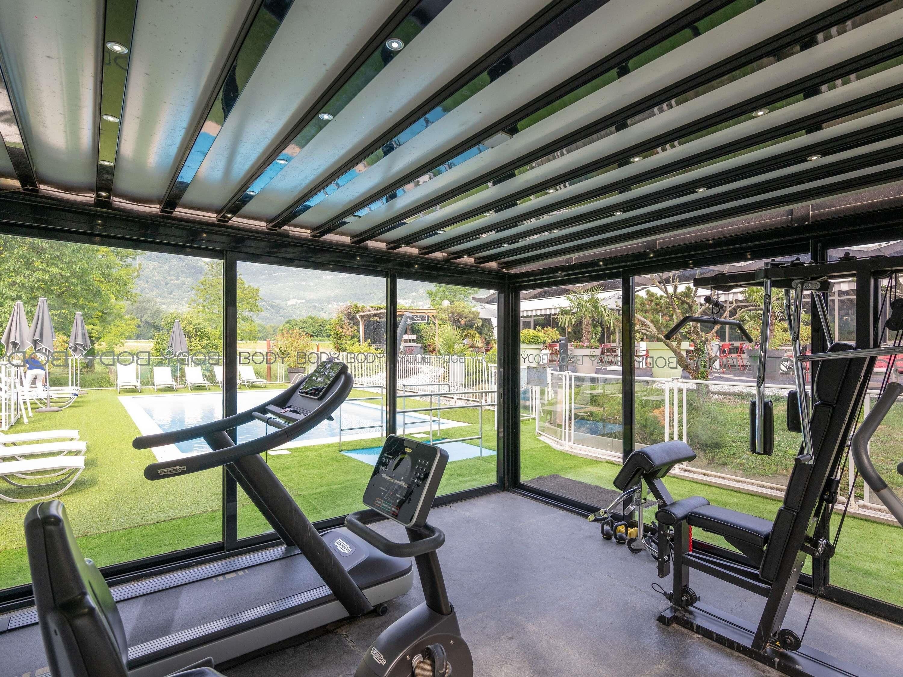 A gym with a treadmill, stationary bike, and weight machines, overlooking a pool and garden.