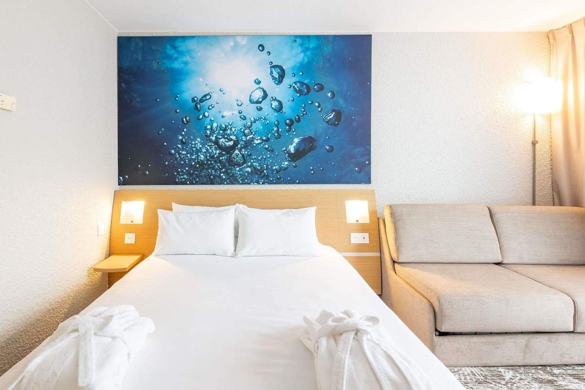 Een hotelkamer met een groot bed, twee bedlampjes, een beige sofa en een schilderij van waterbellen aan de muur.