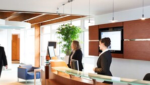 Un espace d’accueil comprenant un comptoir de réception en bois, une grande plante en pot et un écran numérique affichant des informations.