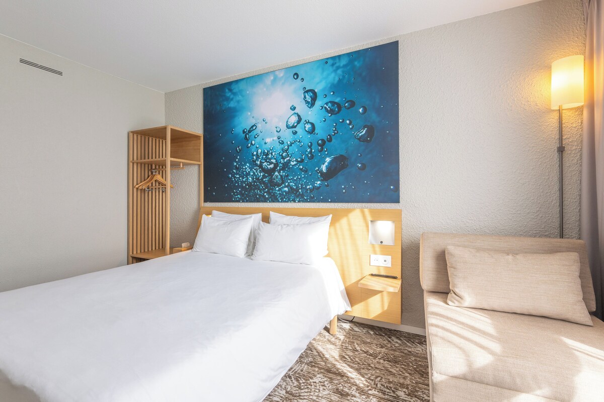Une chambre d’hôtel avec un grand lit, une armoire en bois, une table de chevet avec une lampe et une œuvre d’art fixée au mur représentant des bulles d’eau.