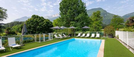 A rectangular swimming pool with a clear blue water surface, surrounded by white lounge chairs and umbrellas, set against a backdrop of lush greenery and mountains.