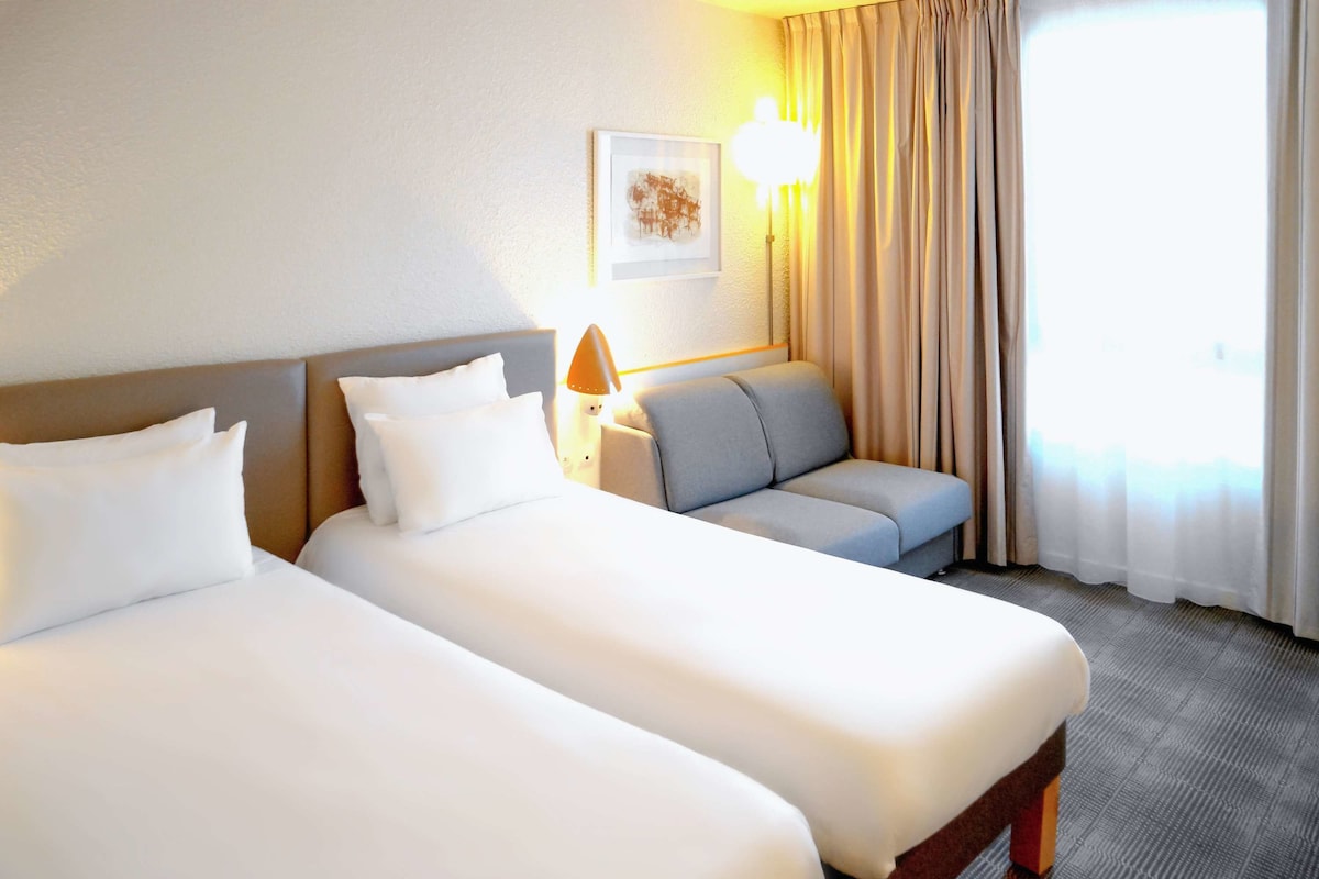 Une chambre d’hôtel avec deux lits, un canapé, une lampe et une fenêtre avec des rideaux.