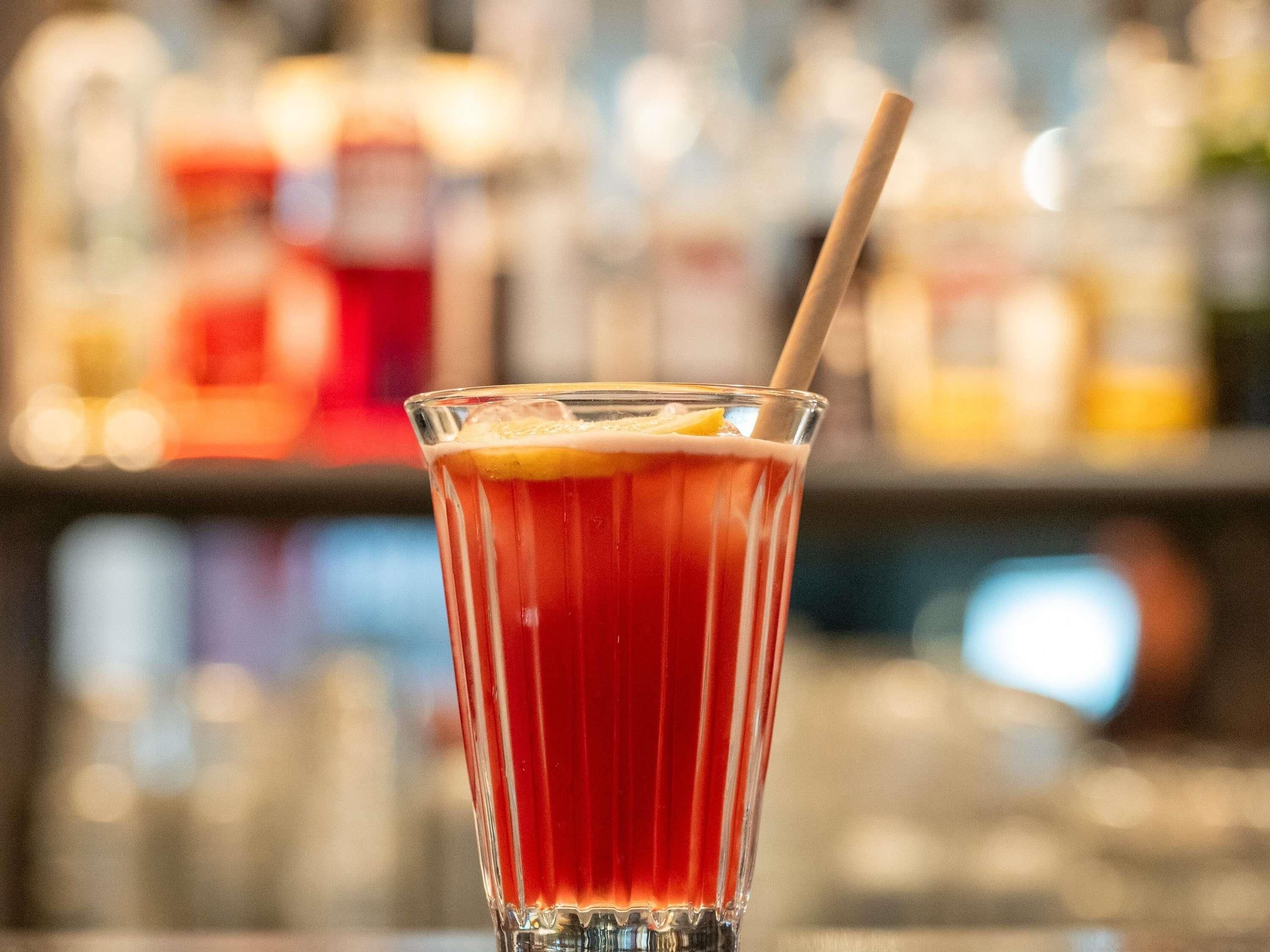 Ein hoher Glas mit Trinkhalm, gefüllt mit einem roten Getränk, vermutlich ein Cocktail, auf einer Bartheke.
