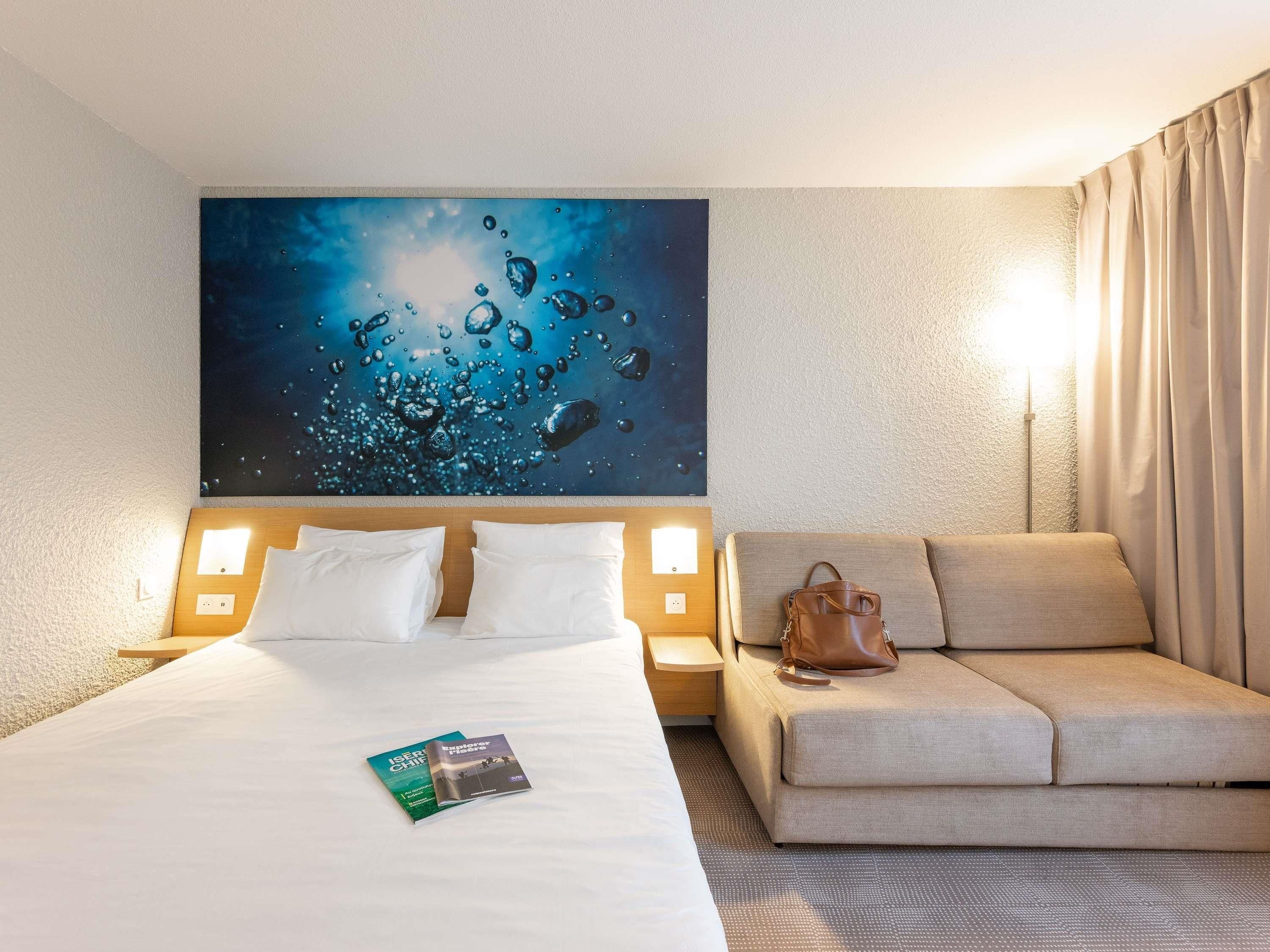 A hotel room with a bed, a sofa, a large wall art of water bubbles, and a book on the bed.