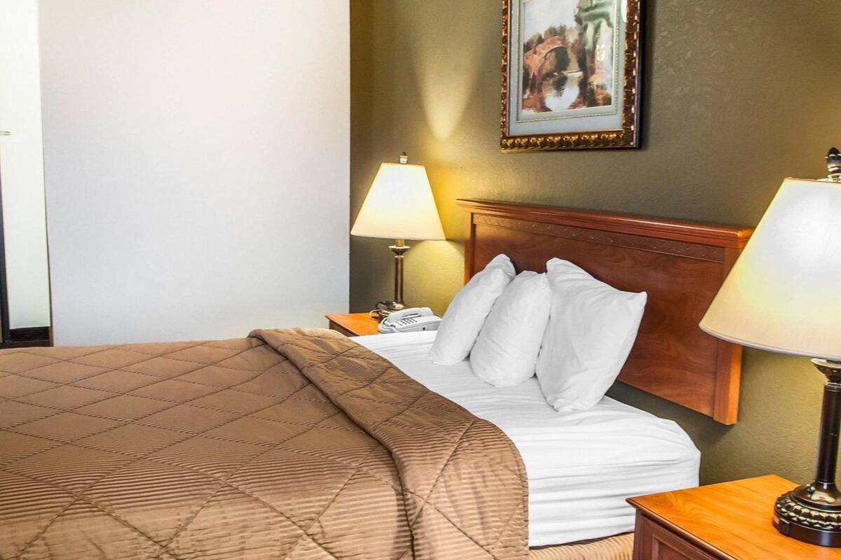 A hotel room with a bed, two bedside lamps, a nightstand, a wooden headboard, and a framed picture on the wall.