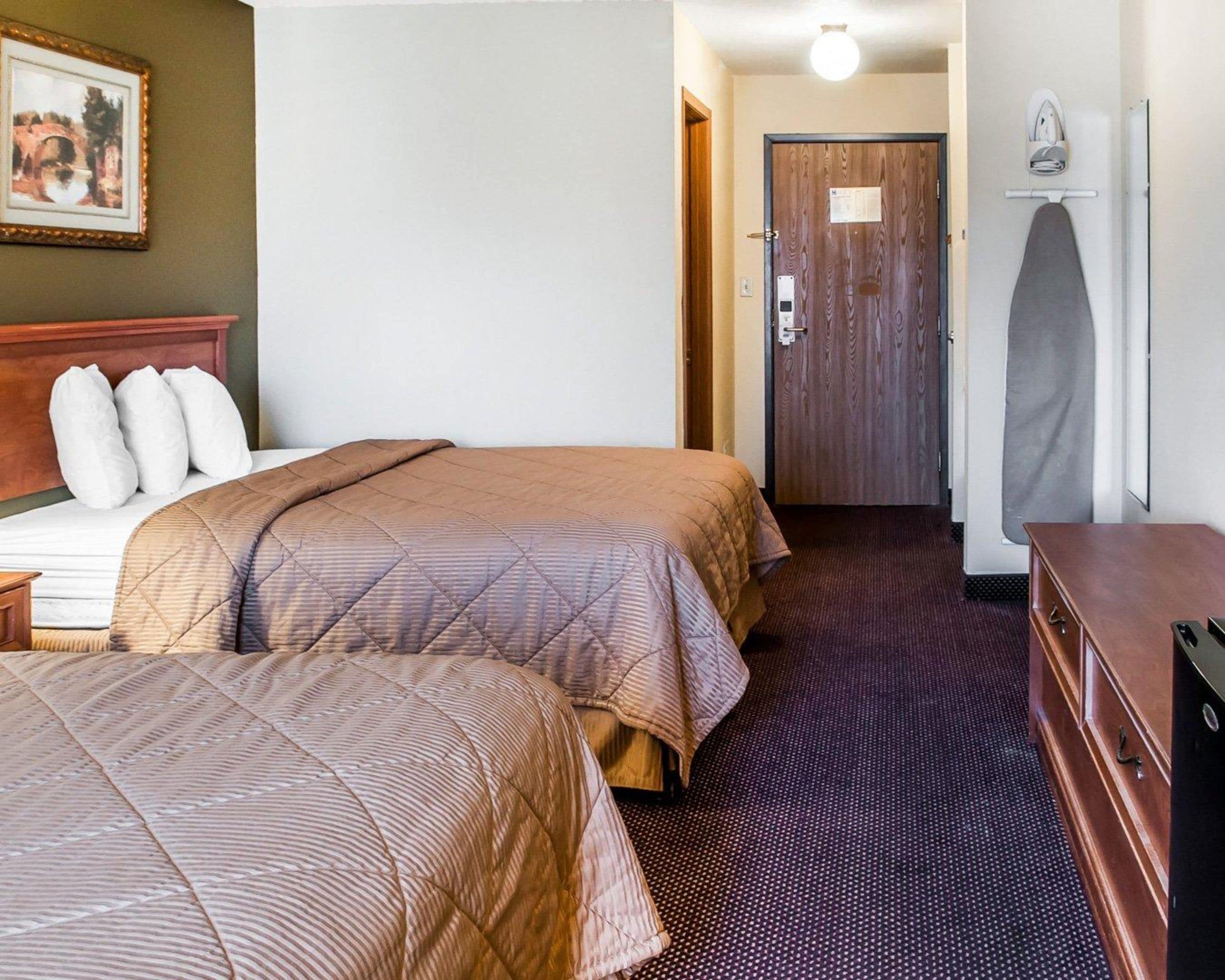 Habitación de hotel con dos camas, un aparador de madera, una puerta con una toalla colgada y un cuadro enmarcado en la pared.