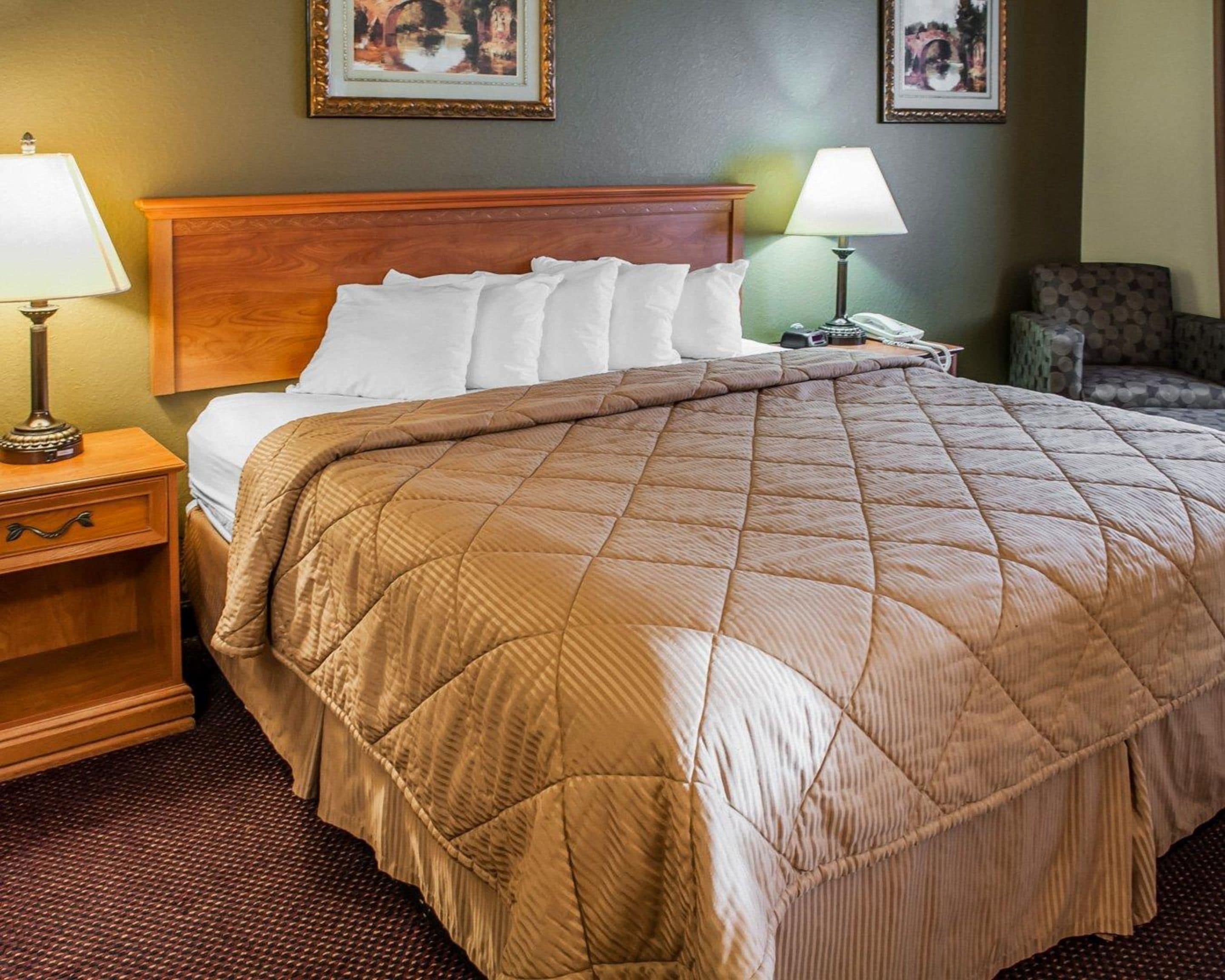 Una cama bien hecha con colcha marrón, cabecera de madera y almohadas blancas. Hay dos mesitas de noche con lámparas y una silla en la esquina.