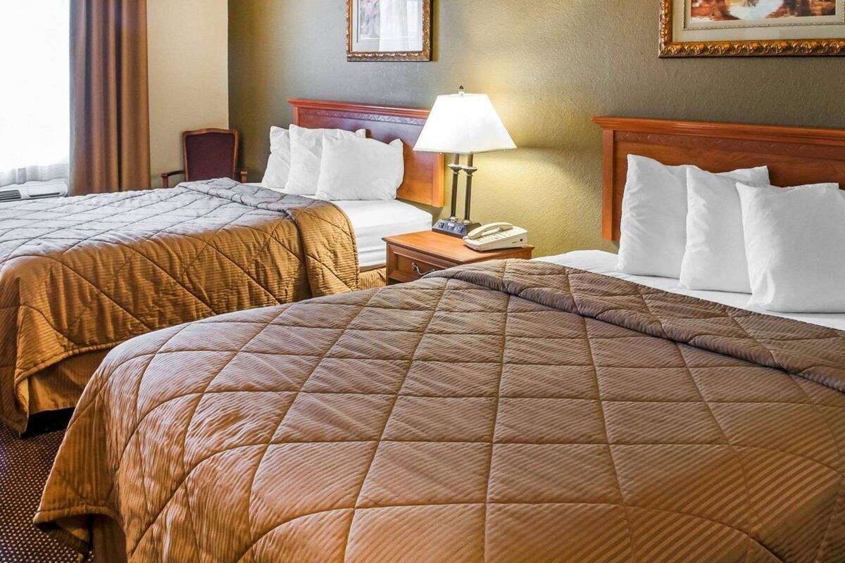 A hotel room with two queen beds, a bedside table with a lamp, a phone, and two framed pictures on the wall.