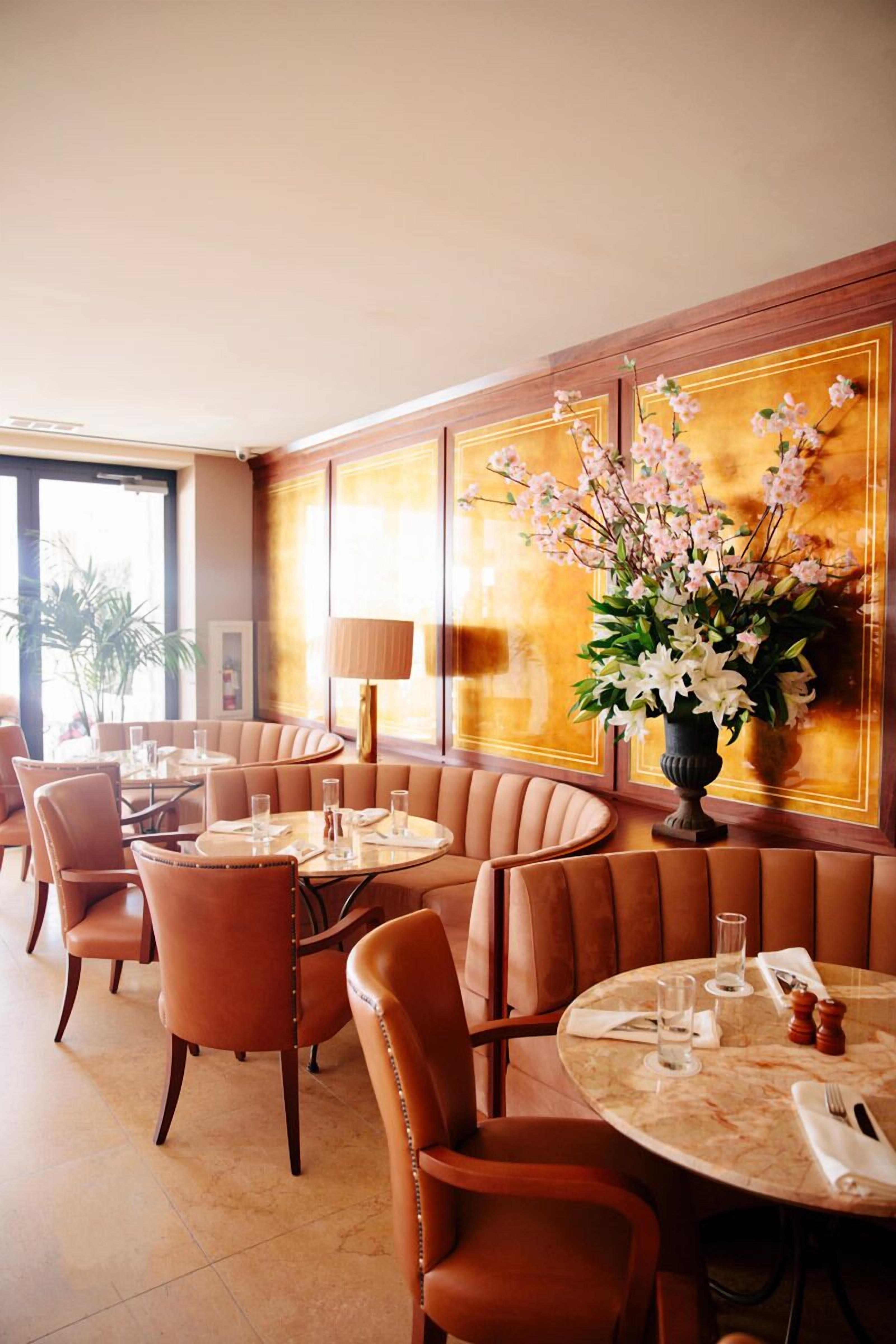 A restaurant with leather booths and round tables, a vase of flowers, and a large window.