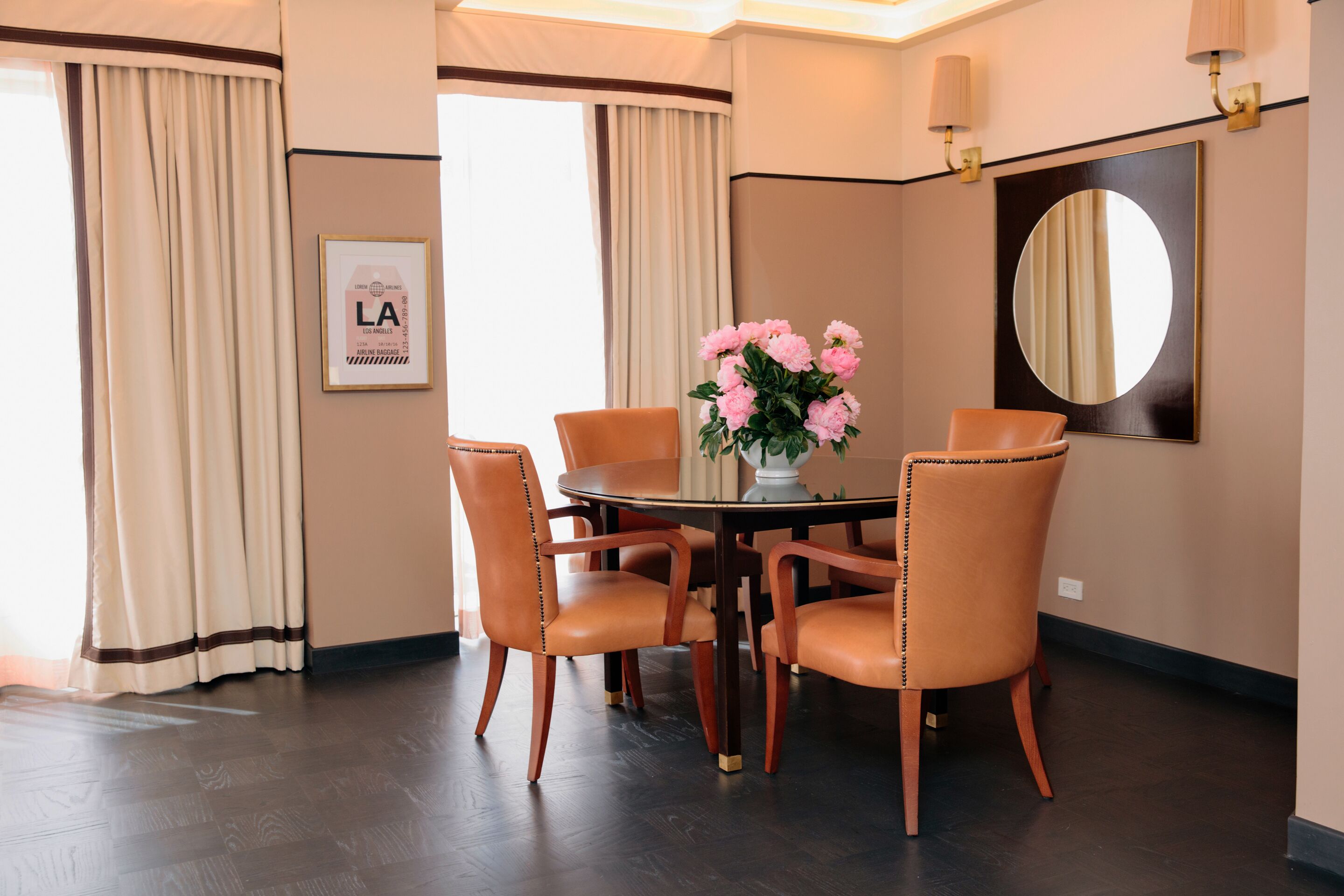 A dining area with a round table, four chairs, a vase of flowers, a mirror, and curtains.
