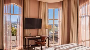 Una habitación de hotel con cama, escritorio con silla, televisor y ventanas grandes con cortinas.