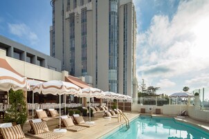 Zona junto a la piscina con sillones de descanso y sombrillas, un edificio alto al fondo y un cielo despejado.