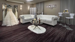 Quarto com um sofá branco, uma mesa de vidro redonda, um espelho e um vestido de noiva.