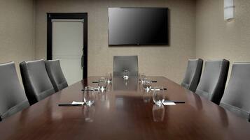 A conference room with a large wooden table, six black leather chairs, a flat-screen TV on the wall, and a door.