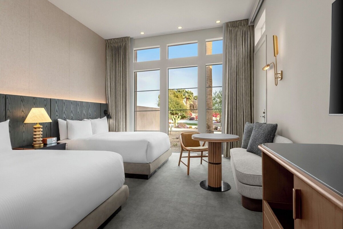 A hotel room with two beds, a small round table, a sofa, a TV, and large windows with a view of the outdoors.