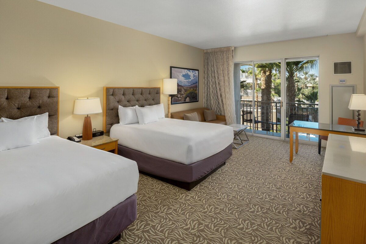 A hotel room with two beds, a desk, a chair, a balcony with a view, and a door.
