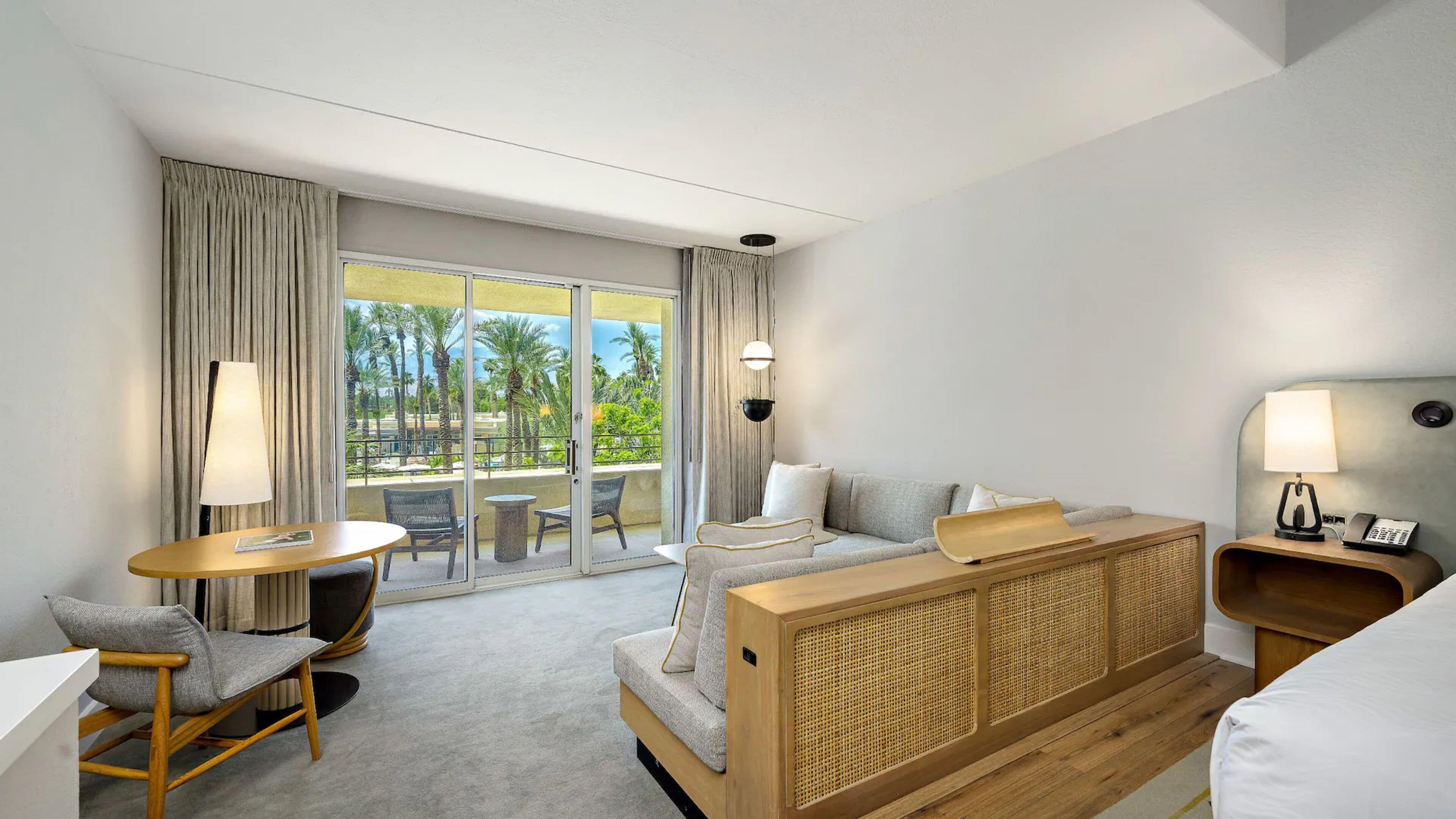 Une chambre d’hôtel avec un canapé, une petite table, une chaise, une table de chevet avec une lampe, et une vue sur les palmiers depuis le balcon.