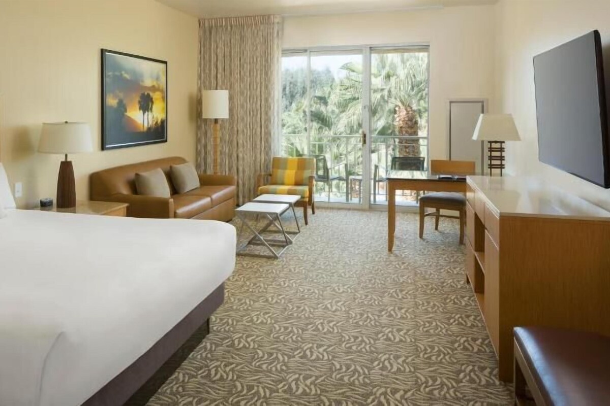 A hotel room with a large bed, a sofa, a desk, a chair, a TV, and a balcony with a view of palm trees.