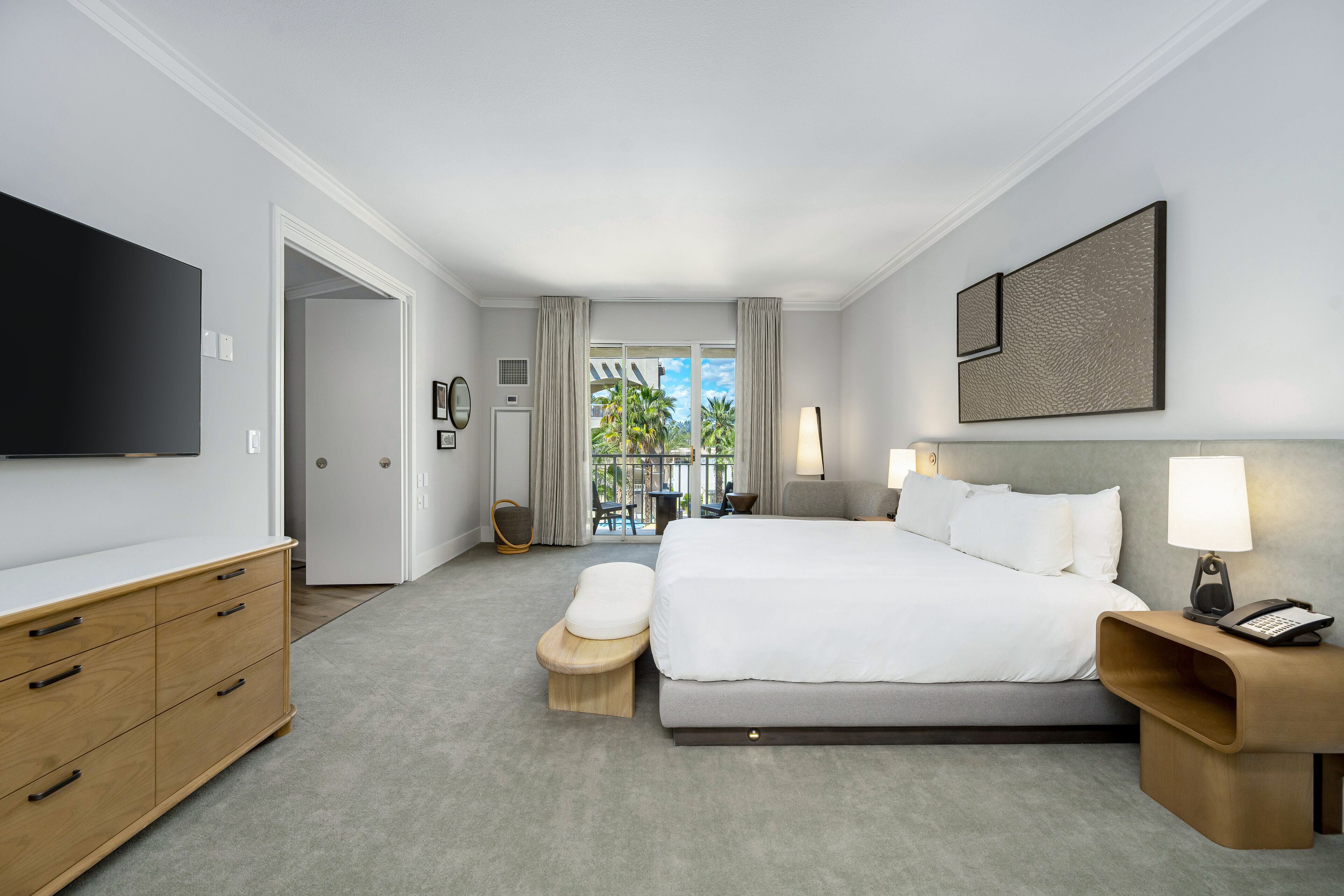 A modern hotel room with a large bed, a flat-screen TV, a wooden dresser, a small seating area, and a balcony with a view of palm trees.