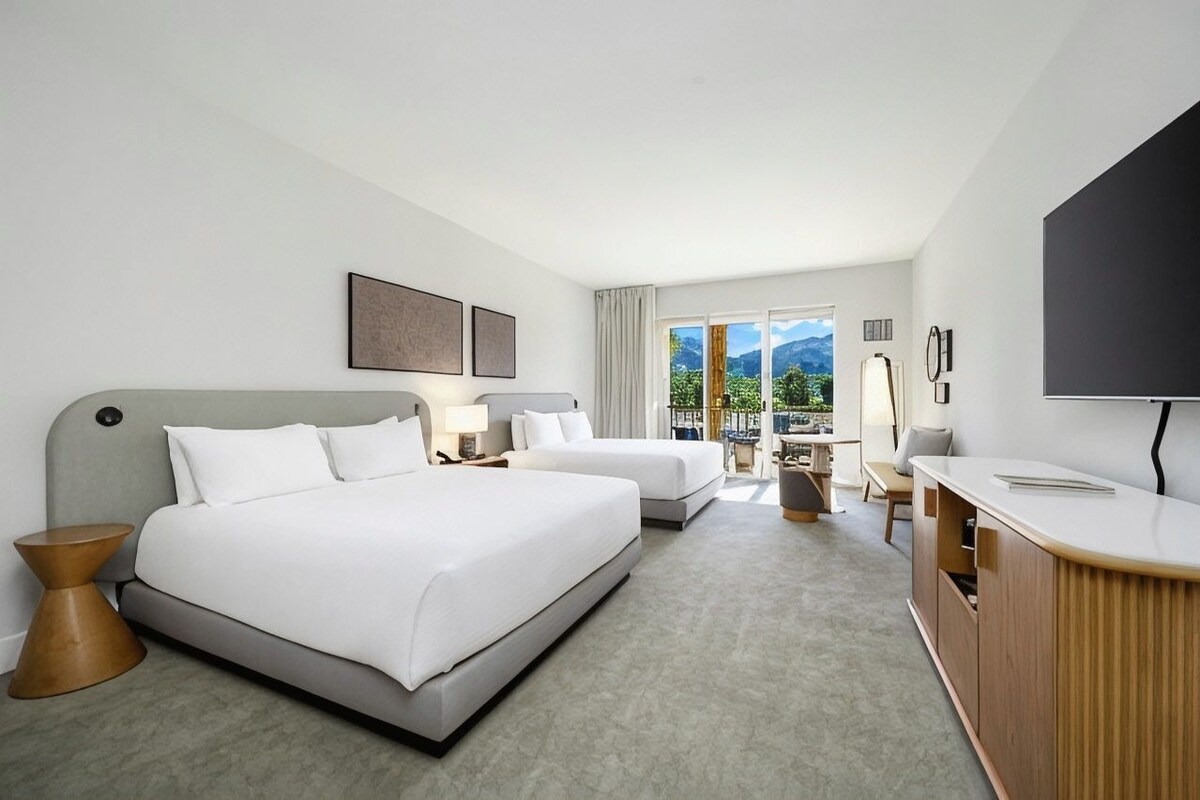 A hotel room with two beds, a desk, a TV, and a view of mountains.