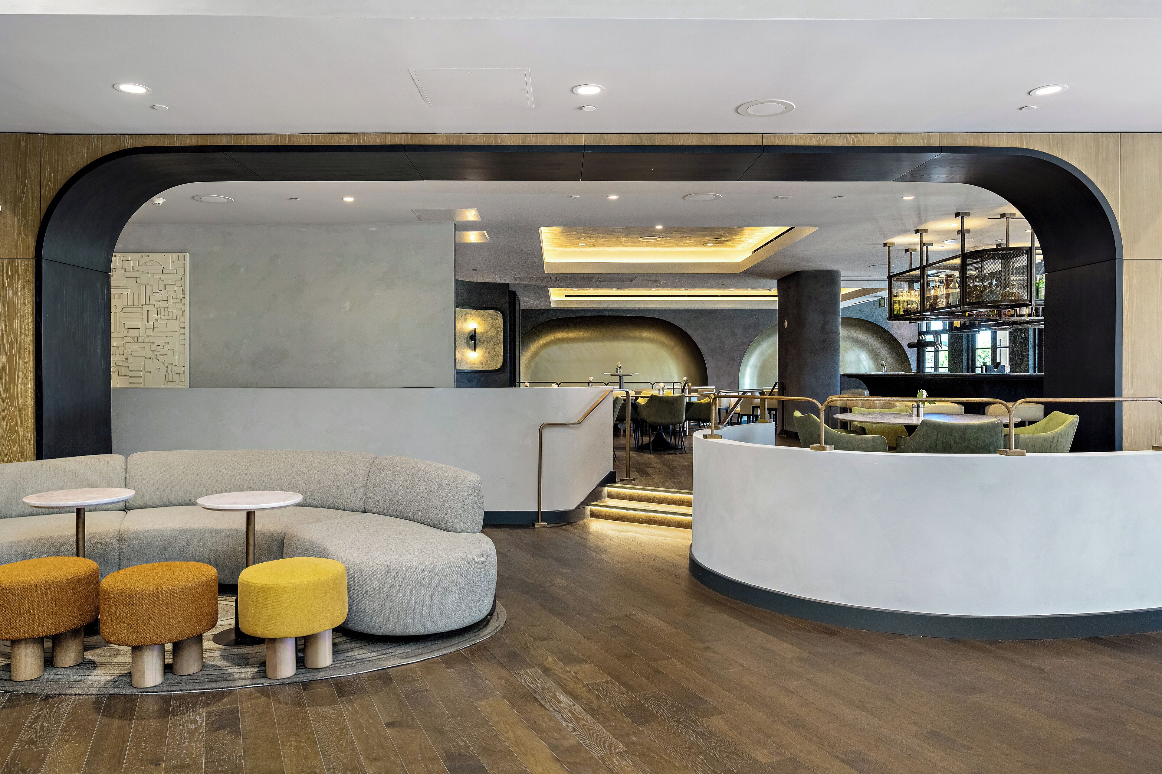 A modern lobby with a curved reception desk, seating area with round tables, and a staircase leading to an upper level.