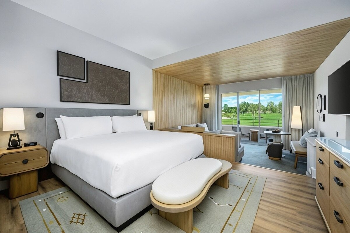 A modern hotel room with a large bed, a desk, a chair, and a view of a green landscape.