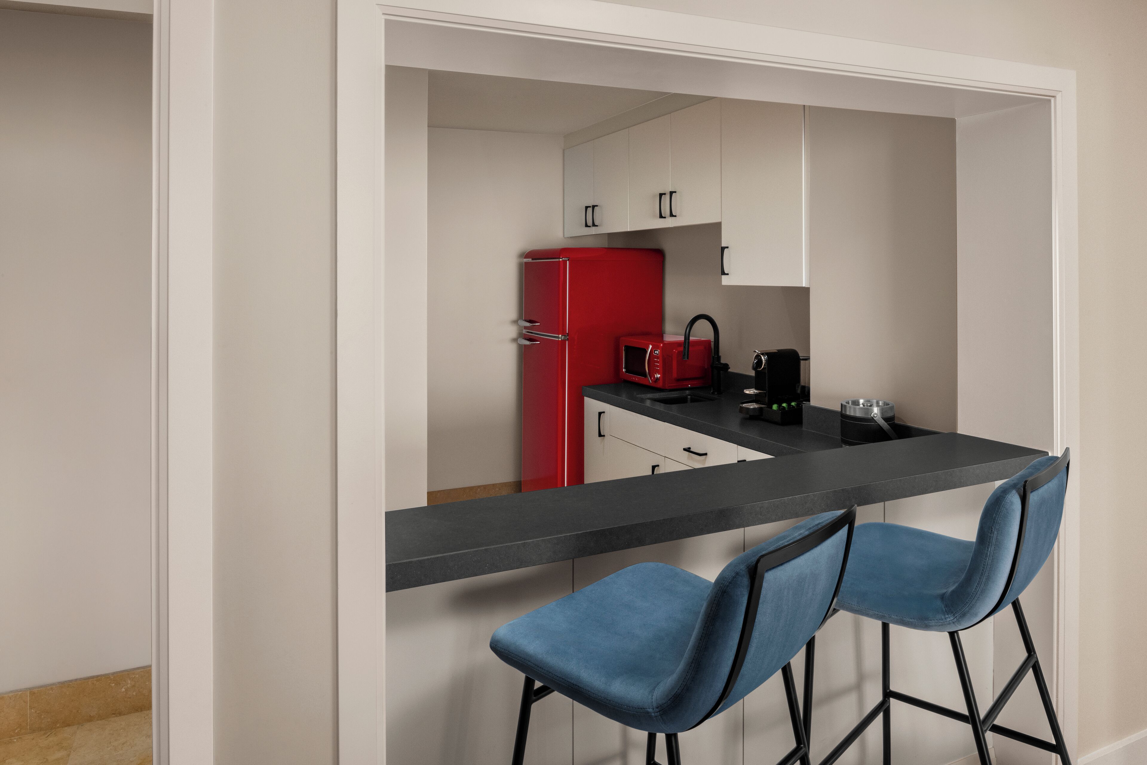 A modern kitchen with white cabinets, a red refrigerator, a black countertop, and two blue bar stools.