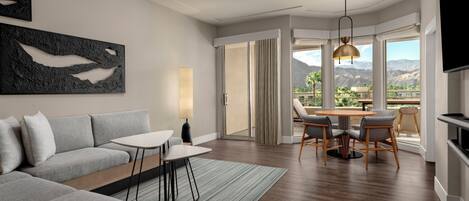 Habitación de hotel moderna con un sofá gris, una pequeña mesa de comedor y sillas. Hay un ventanal con vistas a montañas.