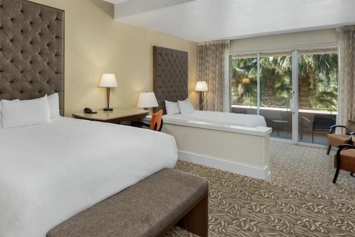 A hotel room with a large bed, a bench, a desk, two lamps, and a view of palm trees through the window.