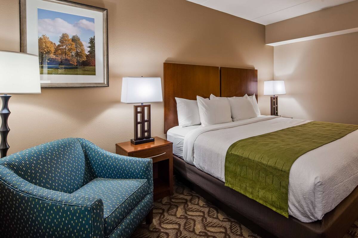 A hotel room with a bed, two bedside lamps, a chair, and a framed picture on the wall.