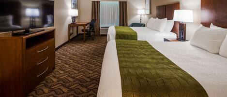 A hotel room with a television, a desk with a chair, two beds with pillows, a lamp, and a patterned carpet.