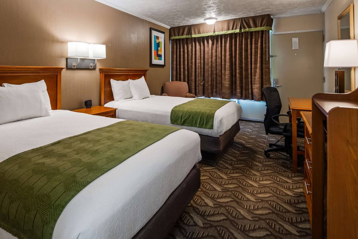 A hotel room with two beds, a desk, a chair, and a lamp.