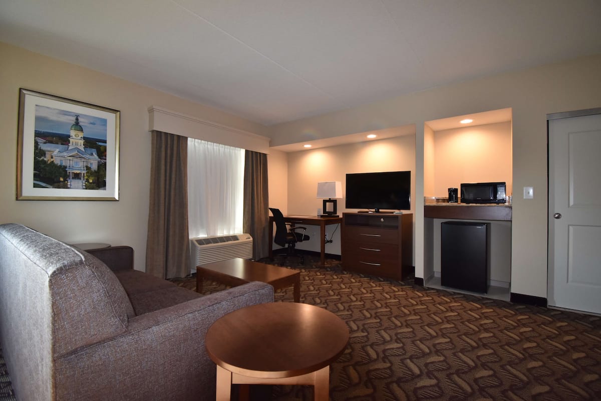 A hotel room with a sofa, a small round table, a desk with a computer and a TV, a mini-fridge, and a door.