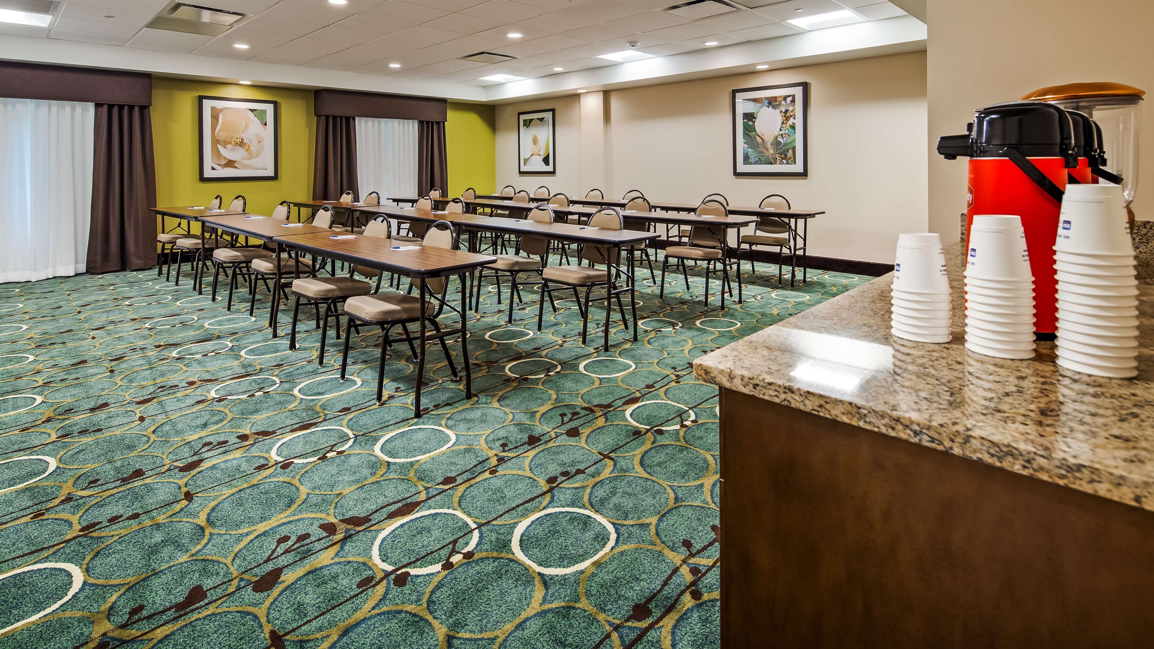 A conference room with a long table, multiple chairs, and a carpeted floor. There are framed pictures on the walls and a coffee machine on the counter.