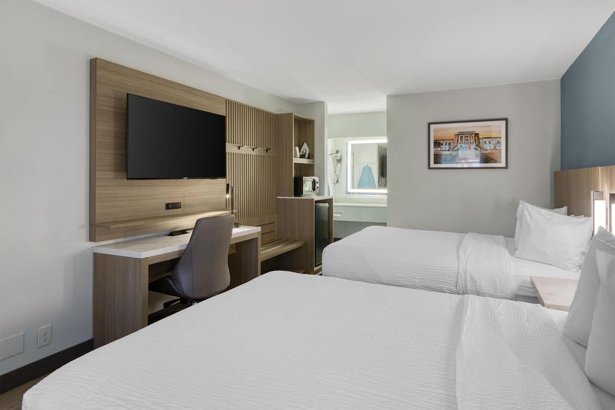 A hotel room with two beds, a desk, a TV, and a bathroom with a shower.