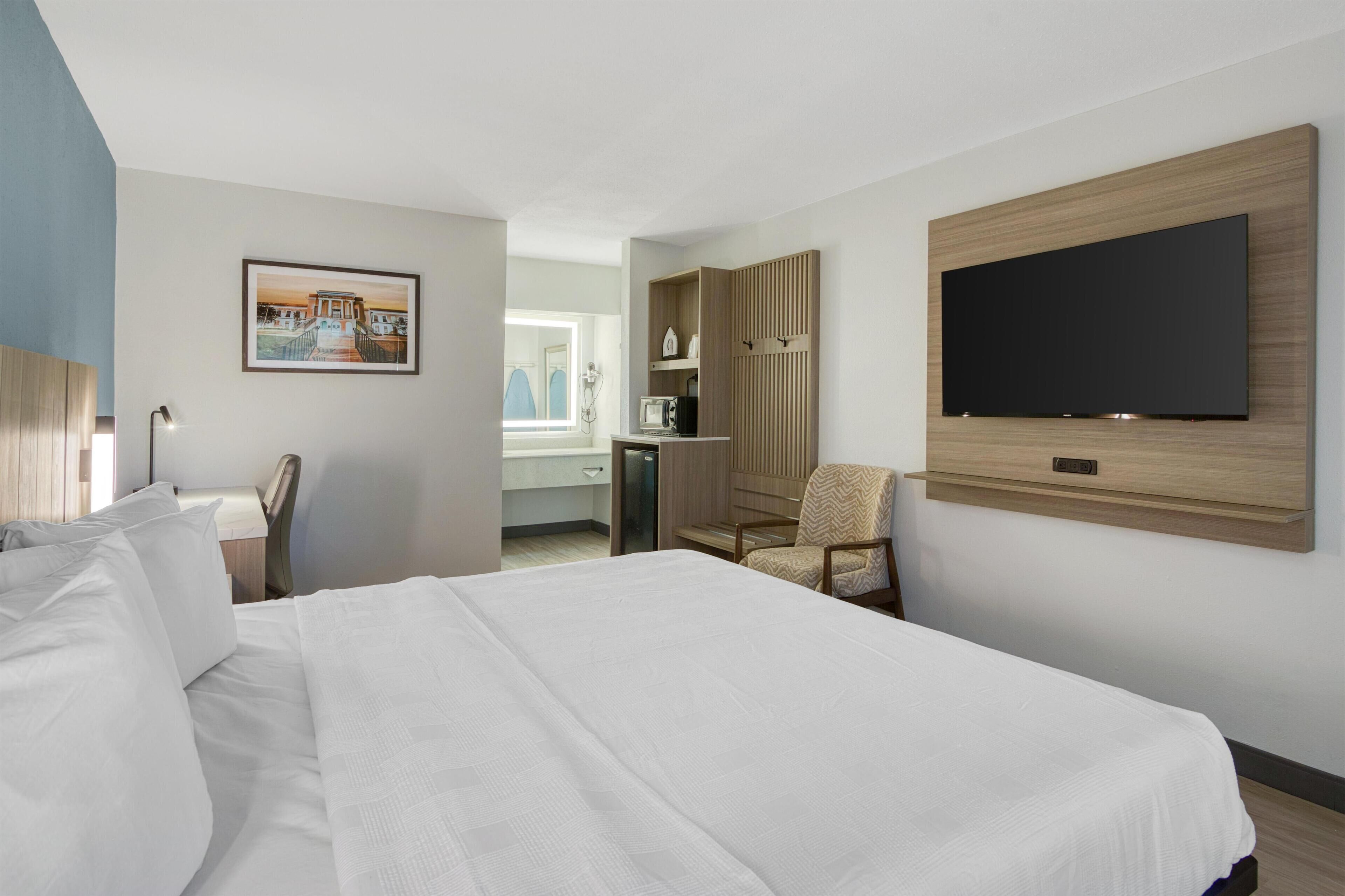 Ein Hotelzimmer mit einem großen Bett, einem an der Wand befestigten Fernseher, einem Sessel, einem Schreibtisch mit Computer und einem Wandbild.