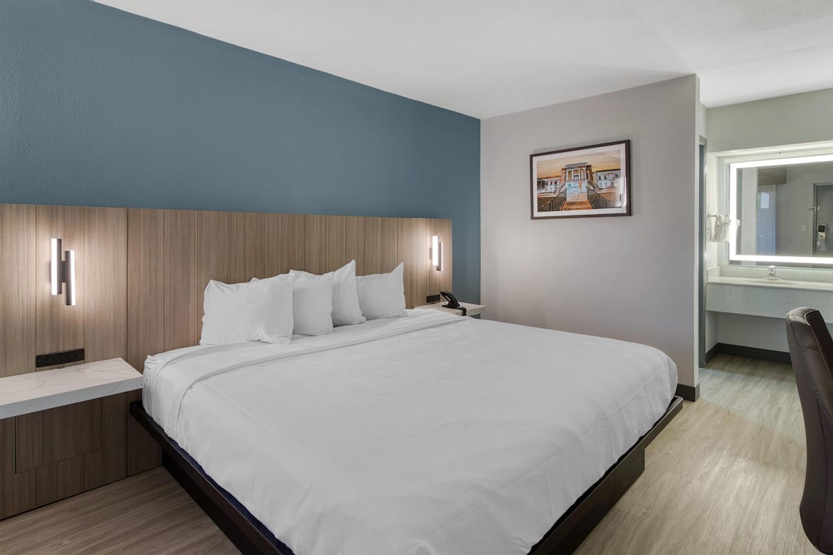 A modern hotel room with a large bed, a wooden headboard, a bedside table, a nightstand, a wall-mounted lamp, a framed picture, and a bathroom with a mirror.