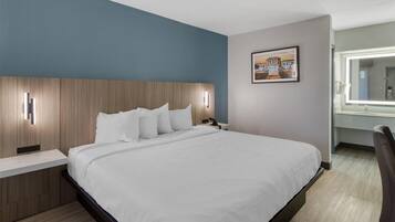 Una habitación de hotel moderna con una cama grande, un cabecero de madera, una mesita de noche, una lámpara de pared, un cuadro enmarcado y un baño con espejo.
