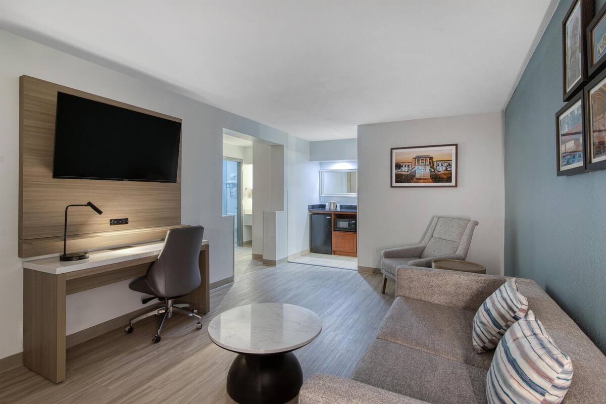 A modern living room with a flat-screen TV, a desk, a chair, a sofa, and a small round table.