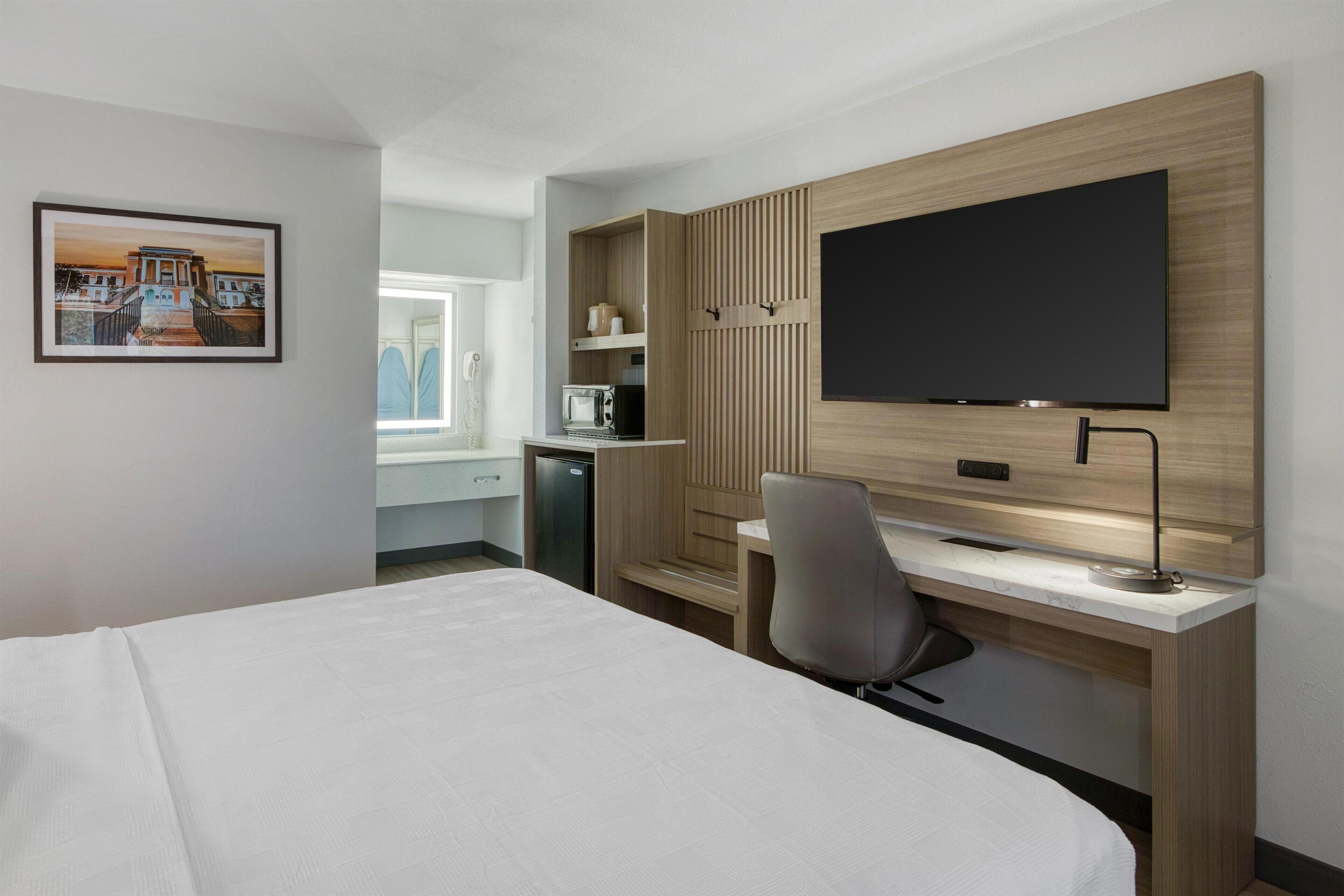 A modern hotel room with a large bed, a desk with a computer, a flat-screen TV mounted on the wall, and a small kitchenette area.