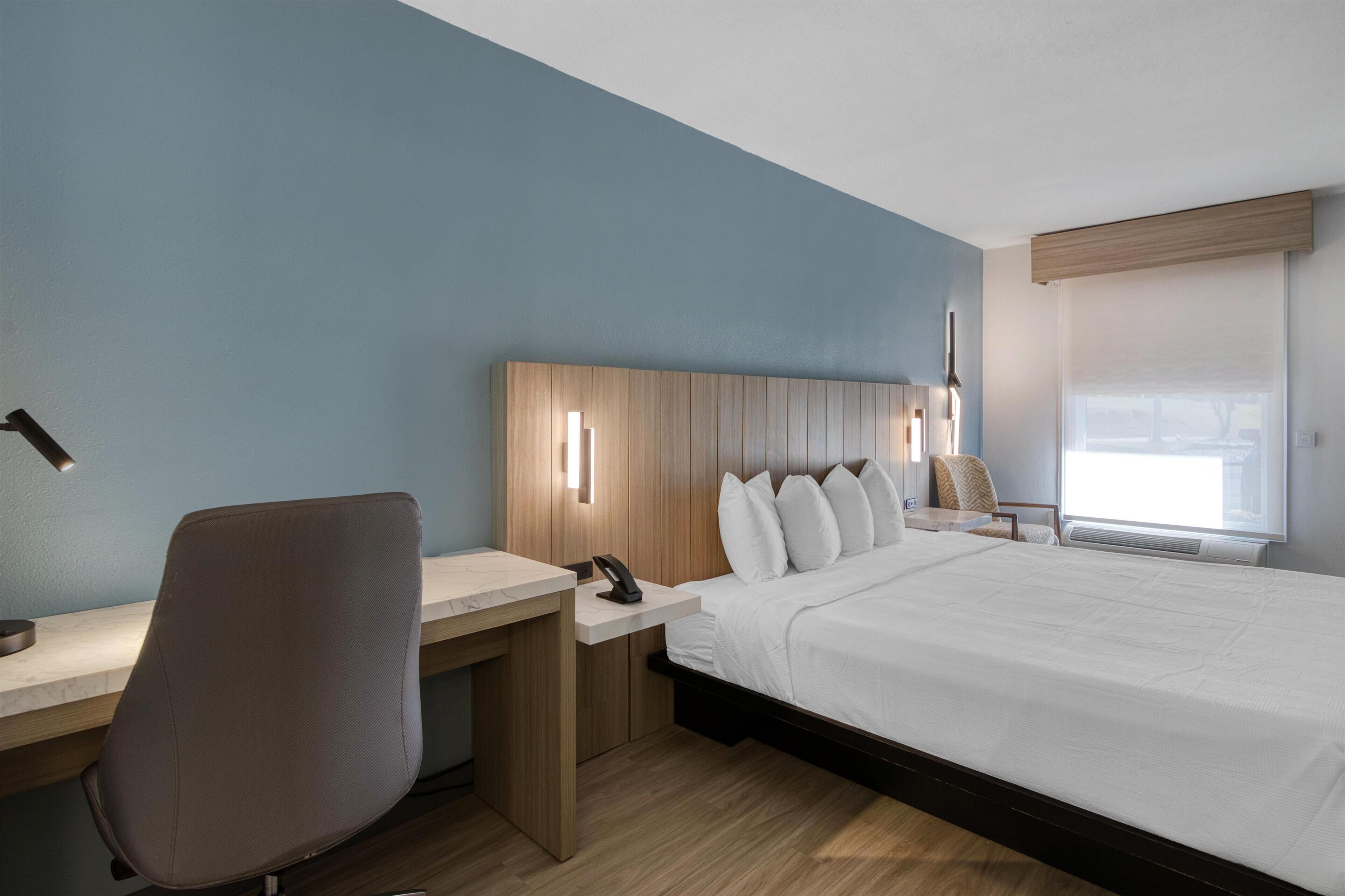 A hotel room with a bed, desk, chair, and a window with blinds.