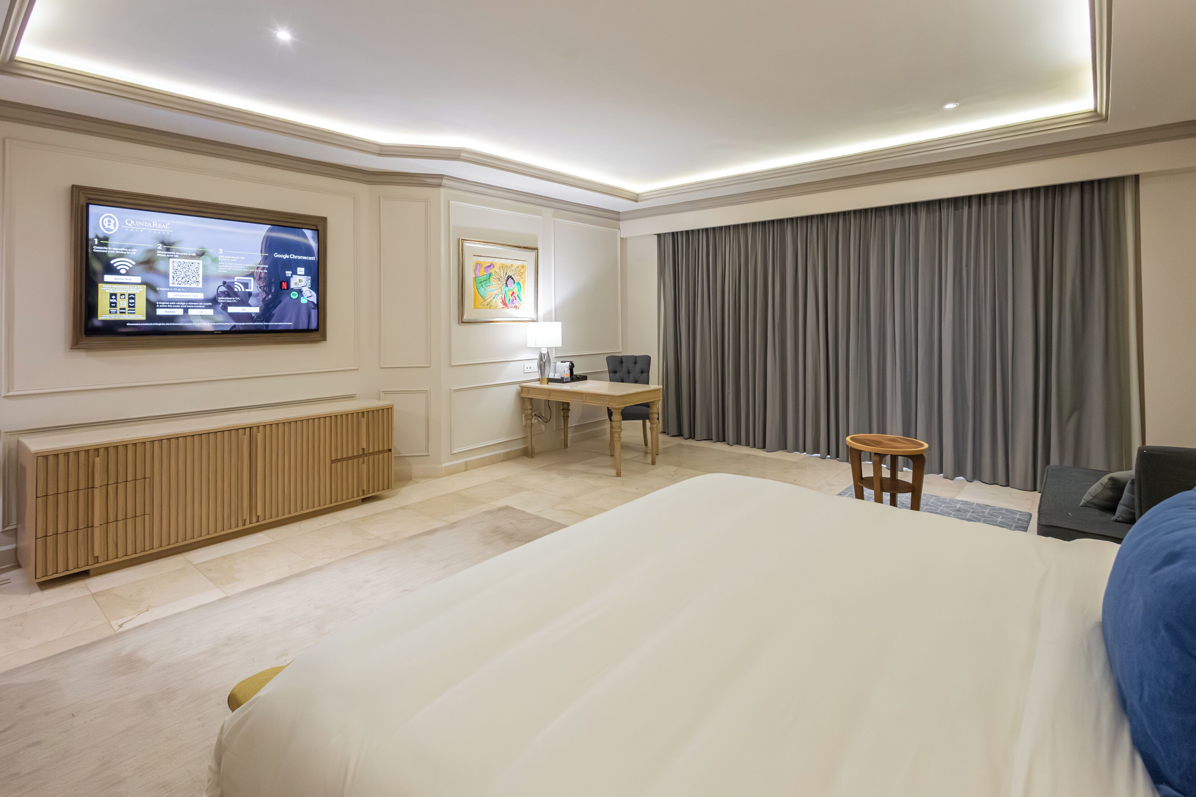 Habitación de hotel con cama, escritorio, silla, televisión y un amplio ventanal con cortinas.