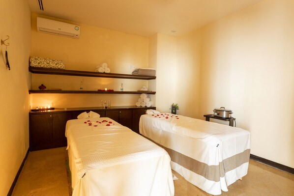 A hotel room with two single beds, a small table, a shelf with toiletries, and an air conditioning unit.