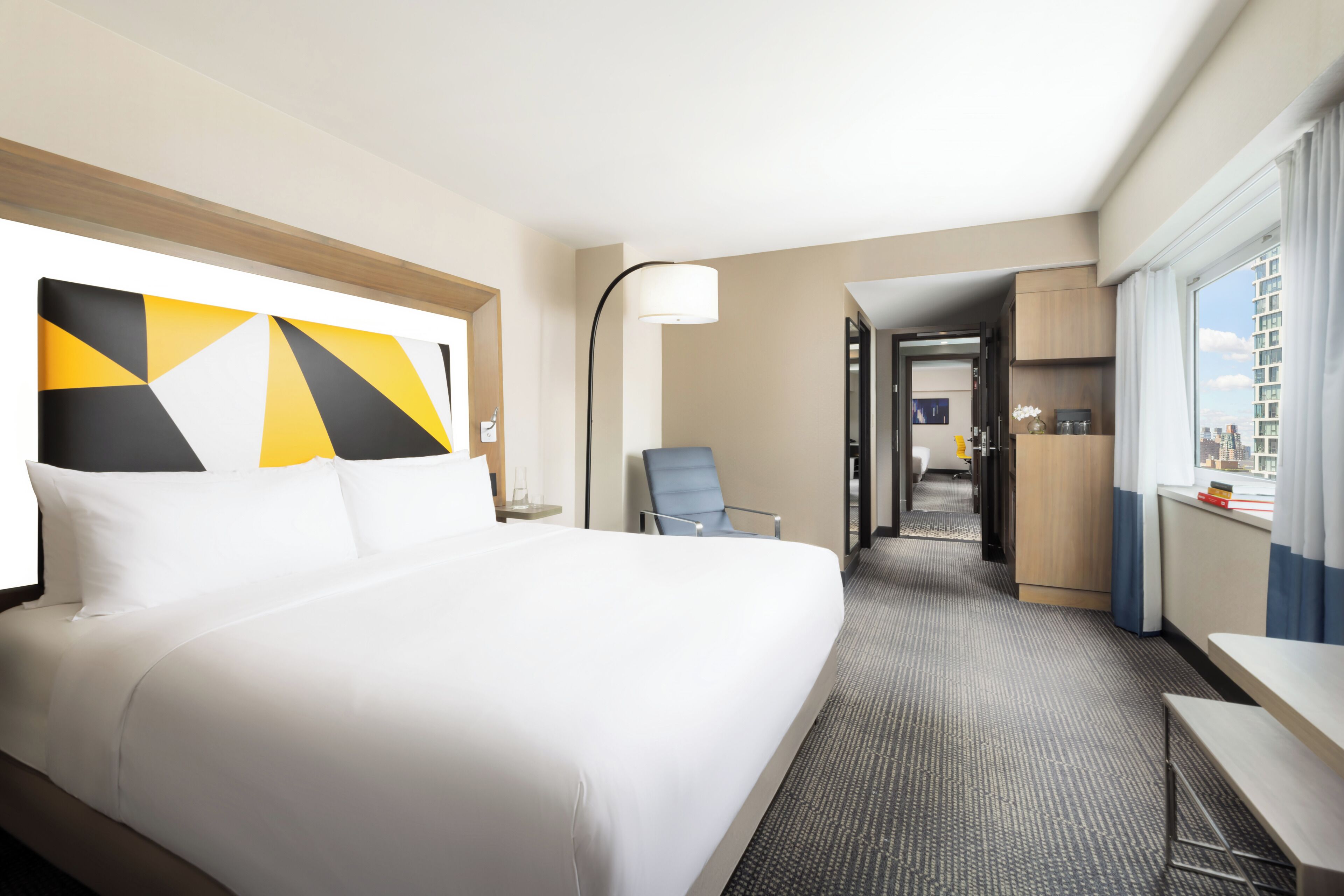 A hotel room with a large bed, a desk, a chair, a lamp, and a view of the city.