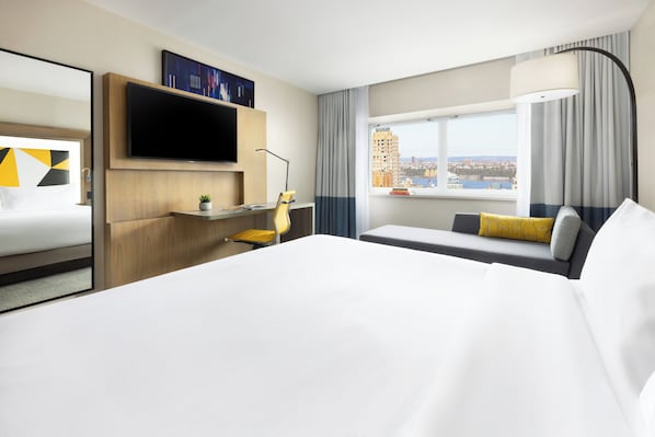 Habitación de hotel con una cama grande, un escritorio con silla, un televisor de pantalla plana en la pared y una ventana con vista a la ciudad.