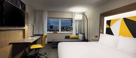 A hotel room with a bed, a desk, a chair, a TV, a window with a city view, and a lamp.