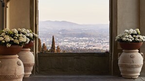 Un arco de entrada con vistas a una ciudad y montañas a lo lejos. A ambos lados del arco, dos macetas grandes de terracota con flores blancas.