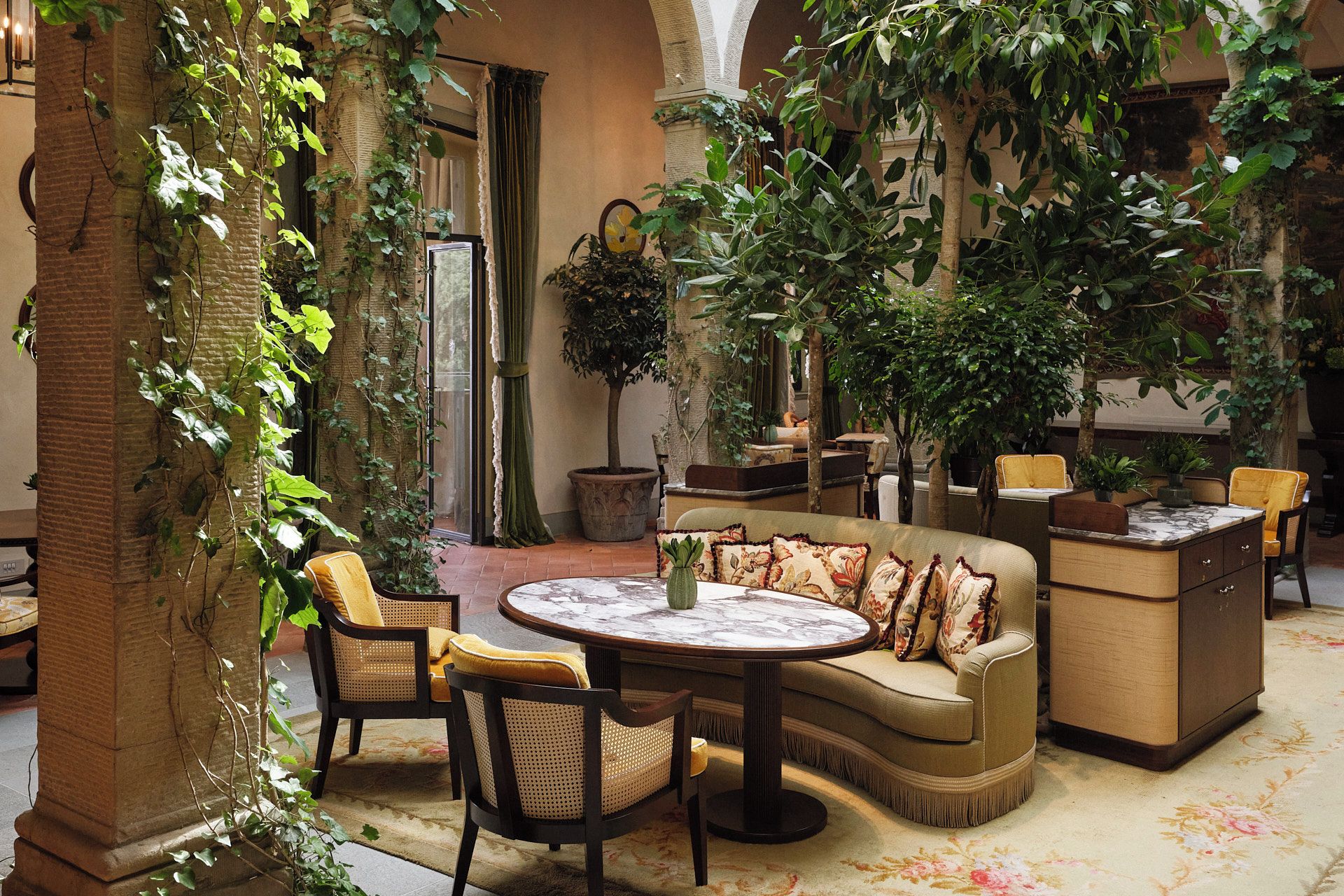 A cozy indoor patio with a round table, cushioned seating, and lush greenery.